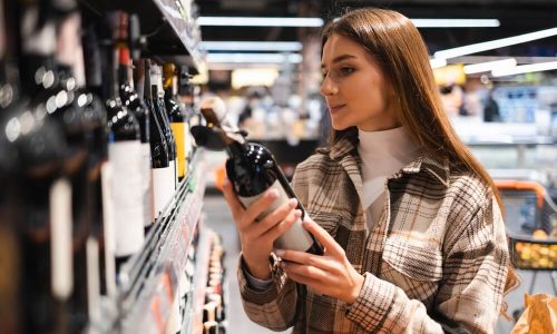 Imagem De Uma Mulher Lendo O Rótulo De Um Vinho Em Um Supermercado.