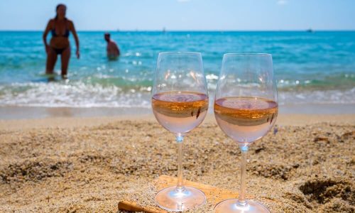 Imagem De Duas Taças De Vinho Rosé Na Praia. Ao Fundo, é Possível Notar O Mar Azul E Pessoas Brincando Na água.