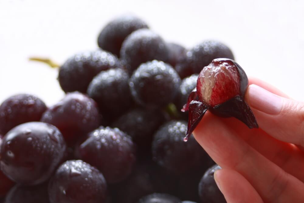 Imagem de uma mão segurando uma uva descascada. Ao fundo, há um cacho de uvas.